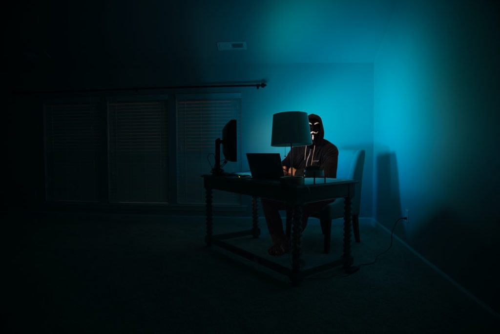person with anonymous mask sitting in front of a laptop in a dark room